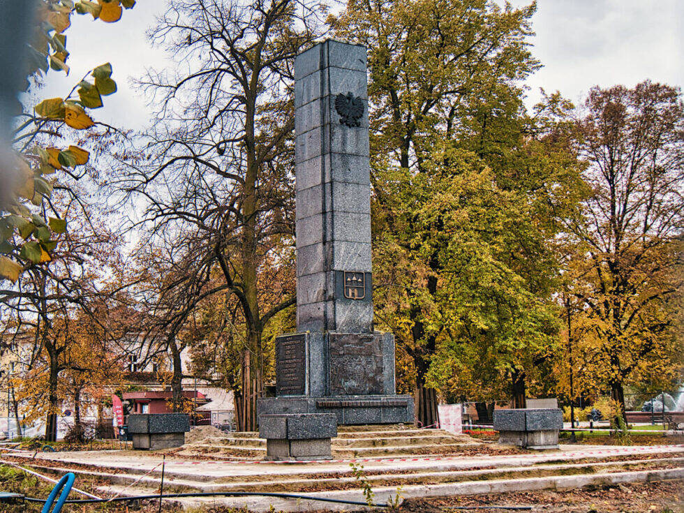 Szary obelisk na postumencie stojący w parku z zamontowanymi tablicami